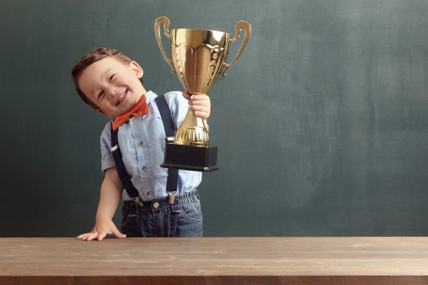 GPT Jongen met een grote glimlach houdt een gouden trofee vast terwijl hij naast een houten tafel staat voor een schoolbord. Hij draagt een lichtblauw overhemd met een oranje vlinderdas en bretels, en een paar donkerblauwe jeans. Zijn vreugde en trots zijn duidelijk zichtbaar.