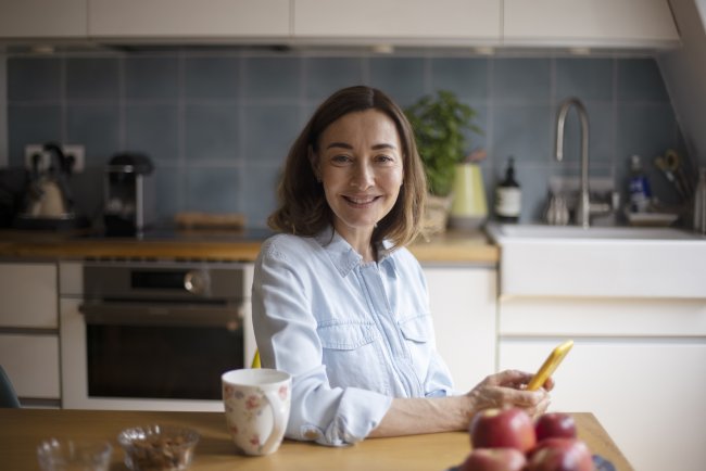 Vrouw zit in de keuken met smartphone, kijkt in camera