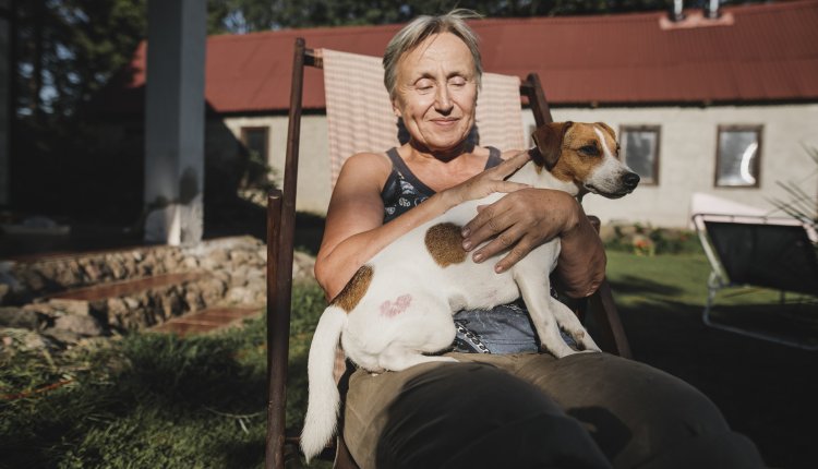 Vrouw zit tevreden op tuinstoel in de zon met hondje op schoot, huis op de achtergrond zichtbaar