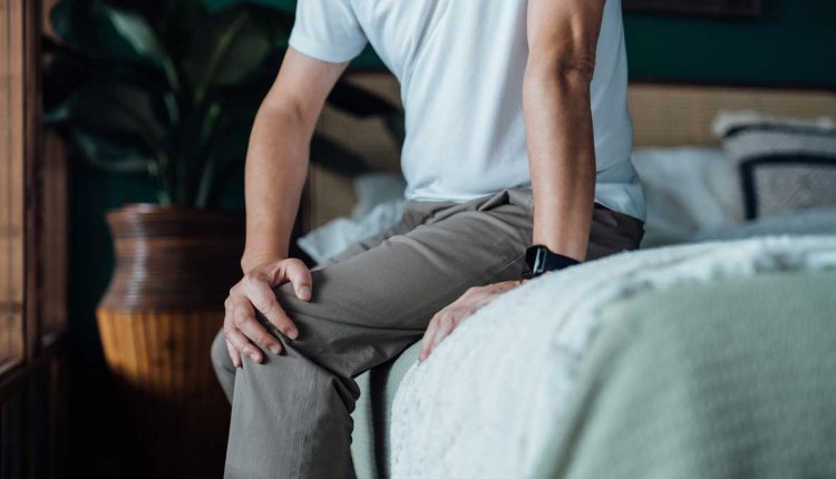 Man met pijnlijke knie zit op bed