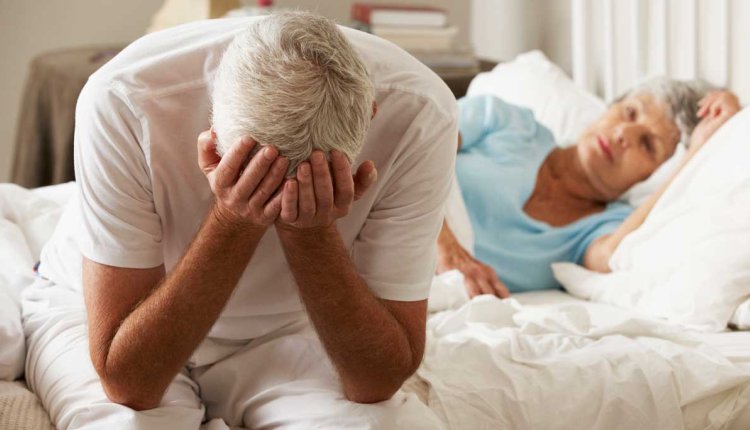 Man met handen voor zijn gezicht in bed met vrouw op de achtergrond