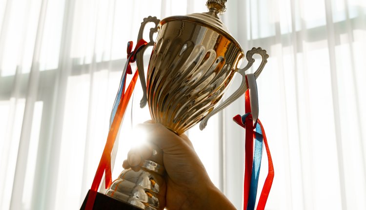 Hand houdt glanzende gouden beker omhoog met rood-blauwe linten, tegen licht raam.