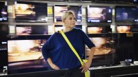 Oudere vrouw is aan het rondkijken in een electronicazaak, veel televisieschermen in beeld