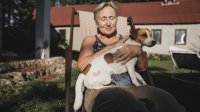 Vrouw zit tevreden op tuinstoel in de zon met hondje op schoot, huis op de achtergrond zichtbaar