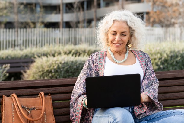 Oudere dame op een bankje buiten lachend bezig op laptop