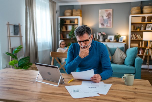 Man is thuis bezig met financien, vrouw op de achtergrond op de bank aan het lezen